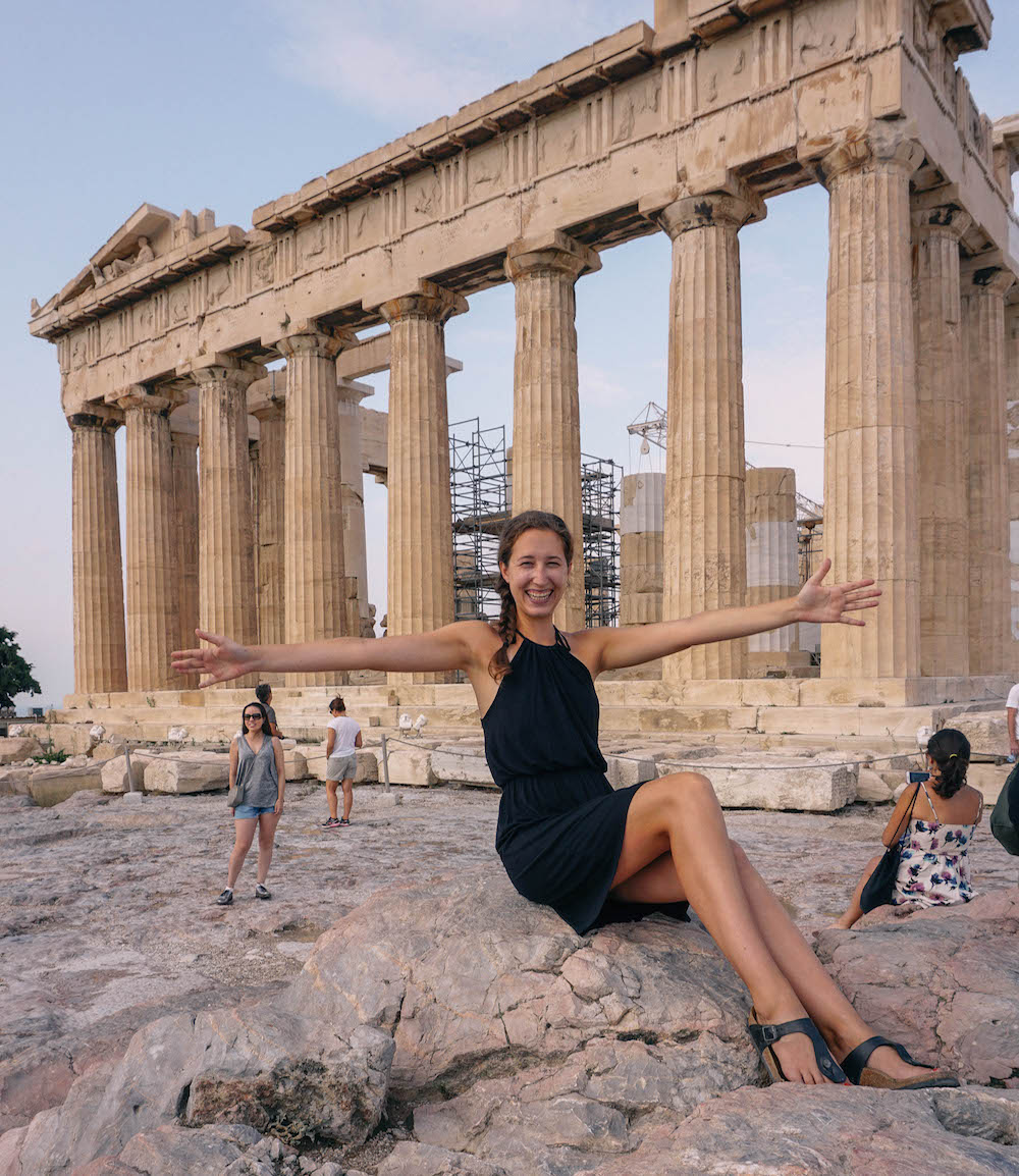 Woman sitting in front of Parthenon, smiling and holding arms outstretched 