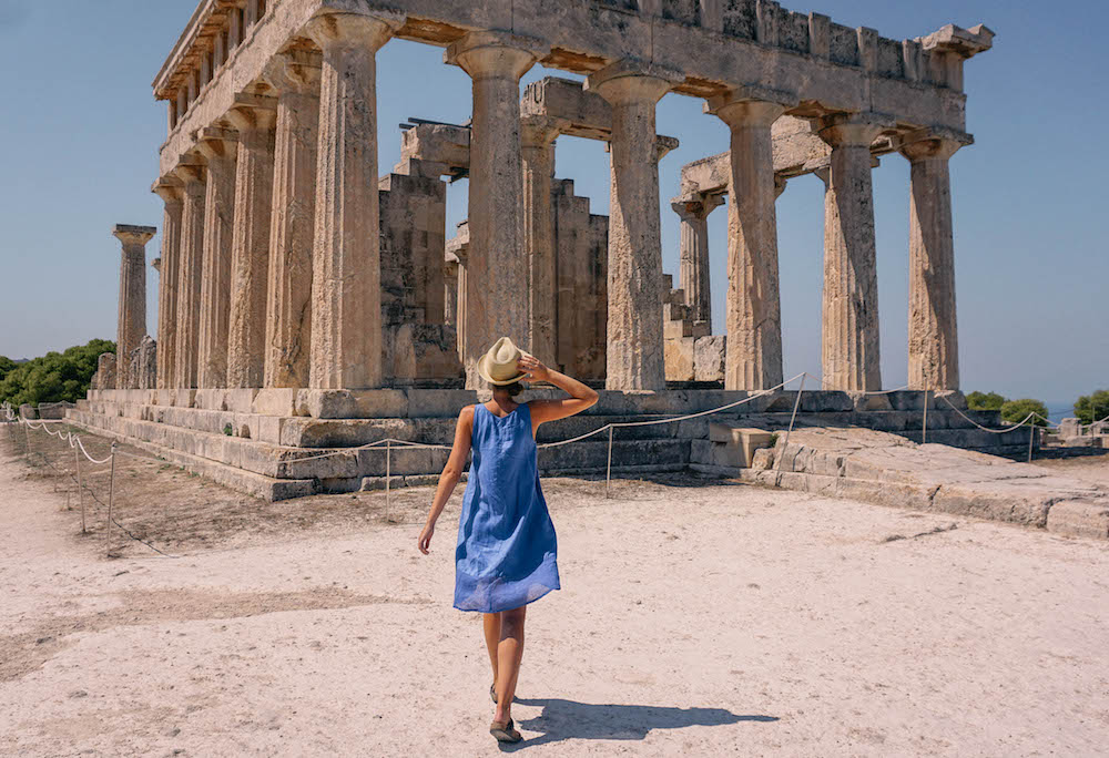 Woman in blue dress walking towards an Ancient Greek temple, with hand on hat. 