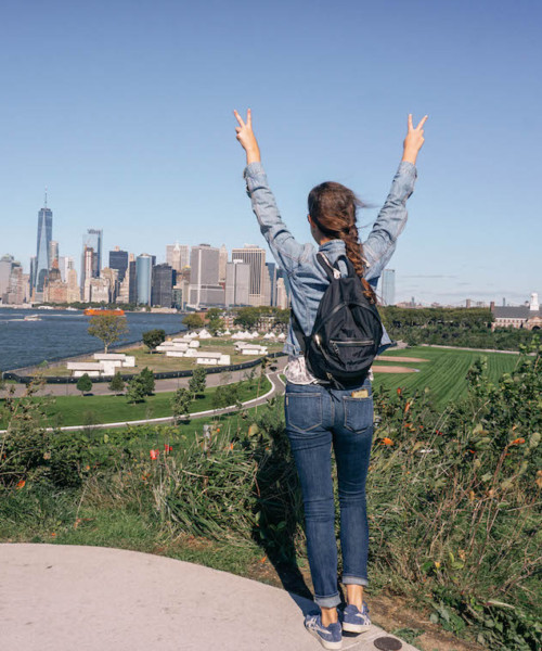 view of woman's back as she holds up peace signs. Manhattan can be seen in the distance.