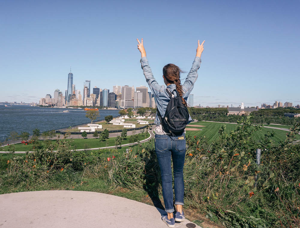 view of woman's back as she holds up peace signs. Manhattan can be seen in the distance. 