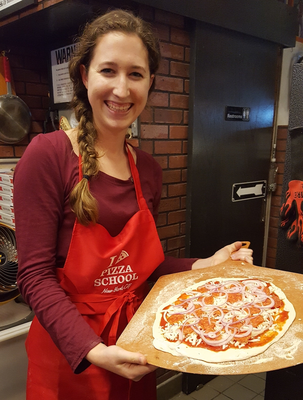 Woman wearing an apron, holding an unbaked pizza aloft. 