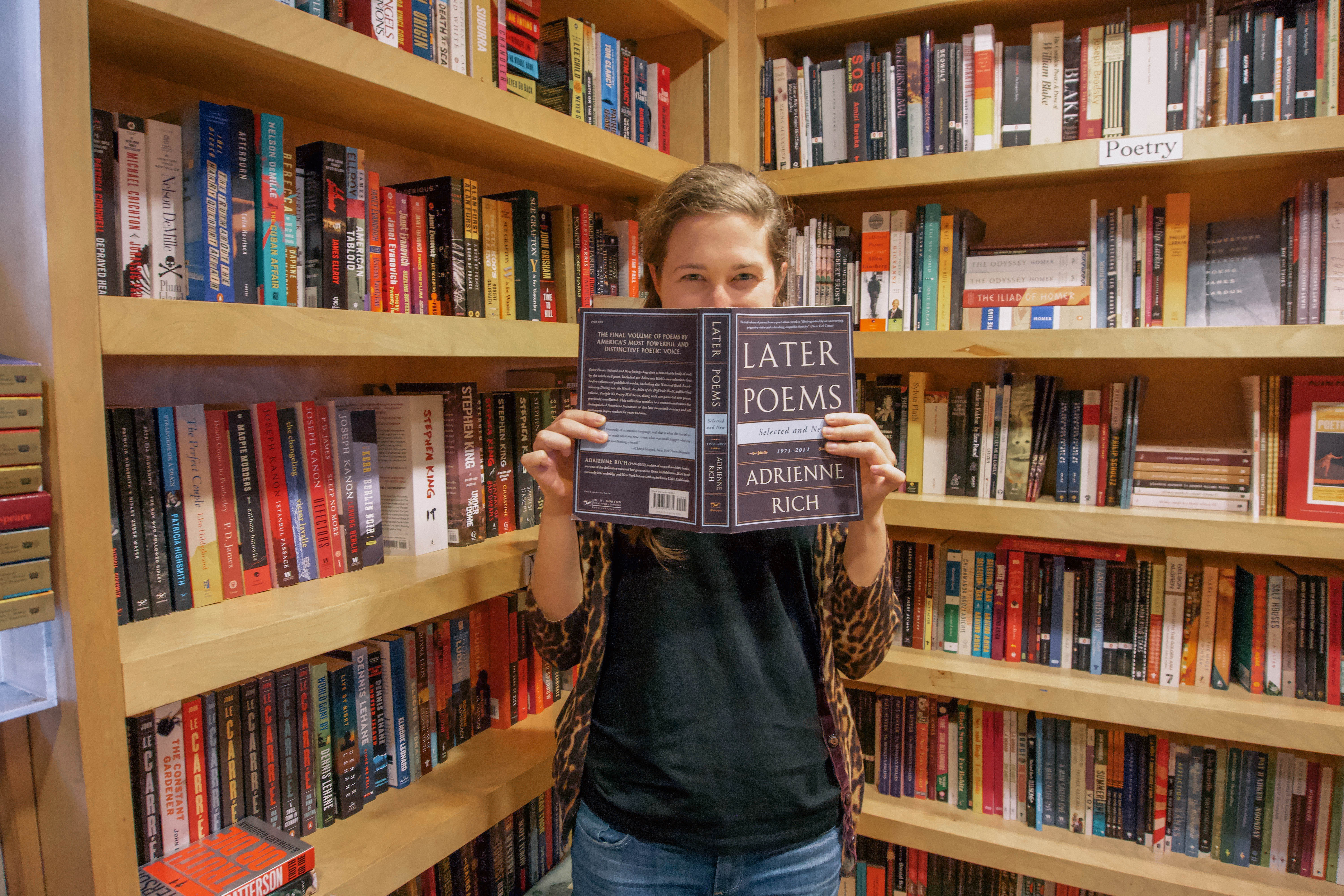 Woman holding an open book in front of her face. She stands between two bookshelves. 