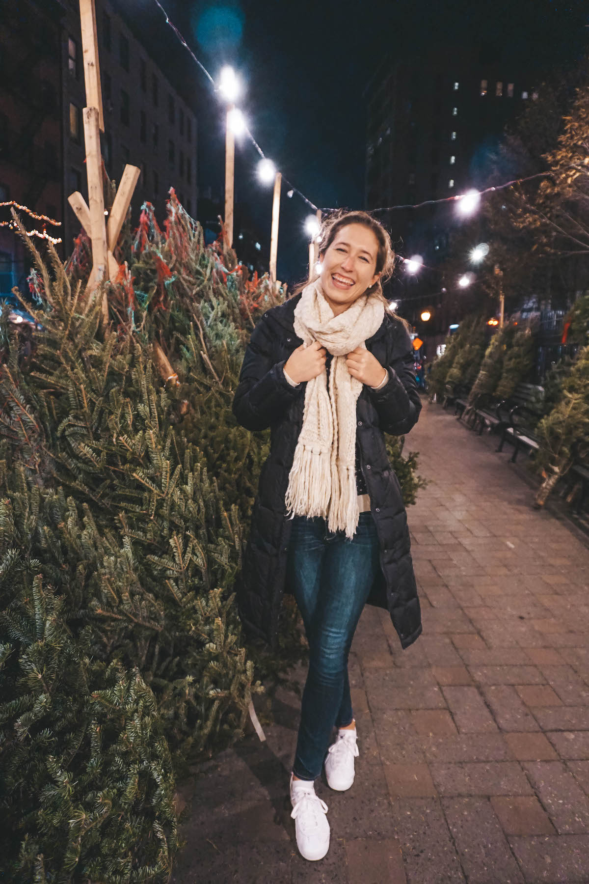 Woman smiling by Christmas trees on sidewalk in NYC. 