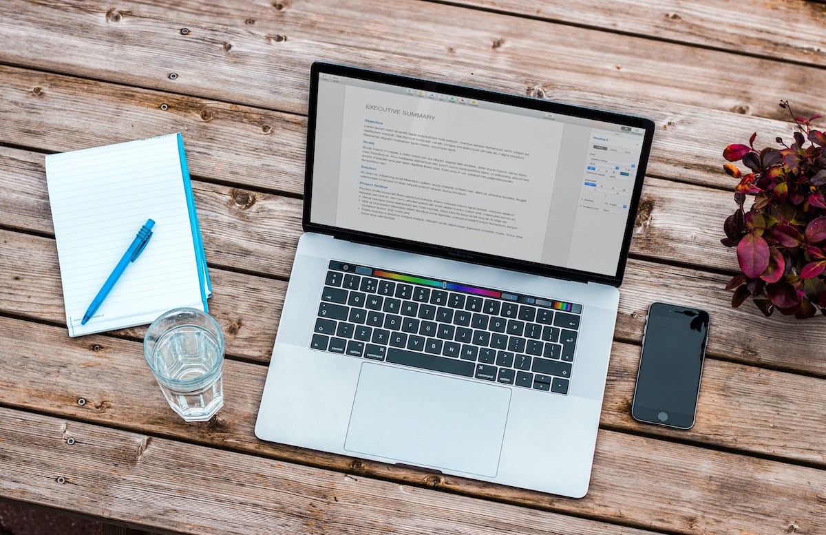 A laptop, glass of water, notebook, and cell phone on a wood table top. 