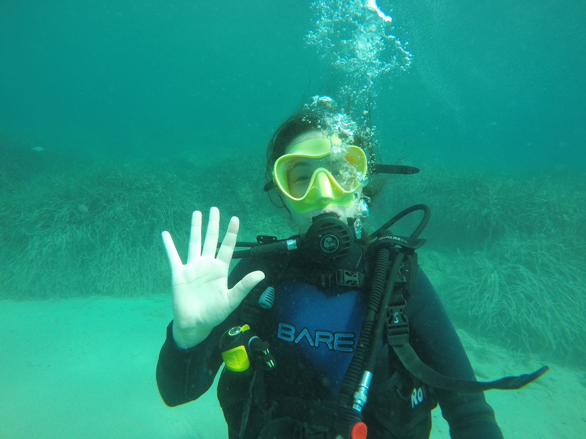 Woman waving under water, wearing Scuba gear. 