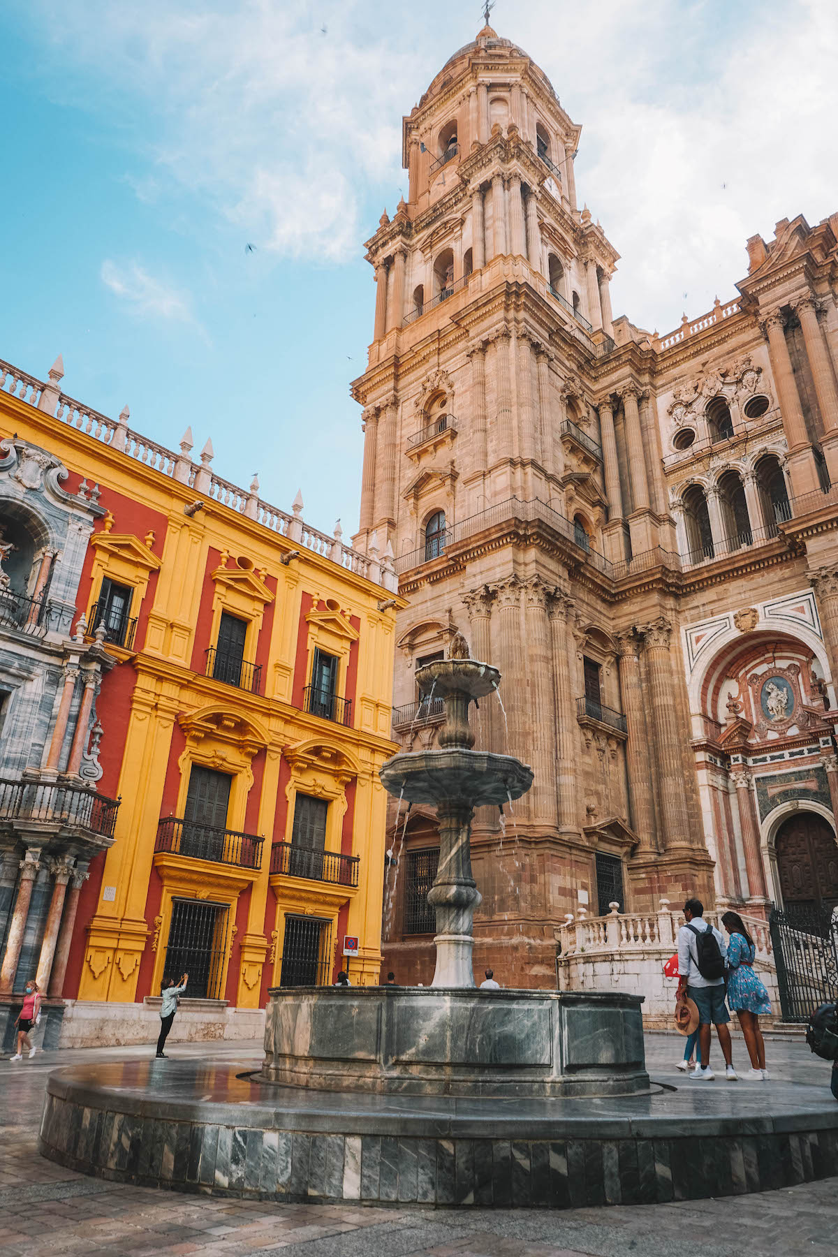 The Malaga Cathedral, seen from a square with a fountain in the middle.